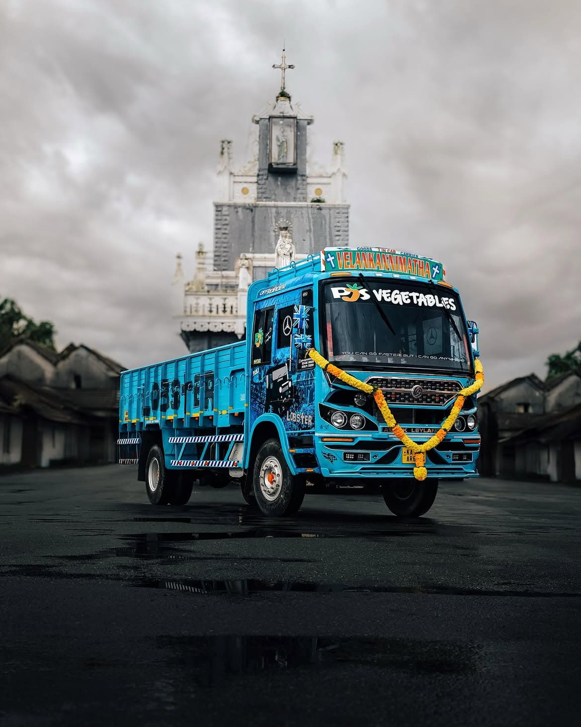 A PJS Vegetables delivery truck, decorated with a marigold garland, parked in front of the Velankanni Matha shrine in Champakulam
